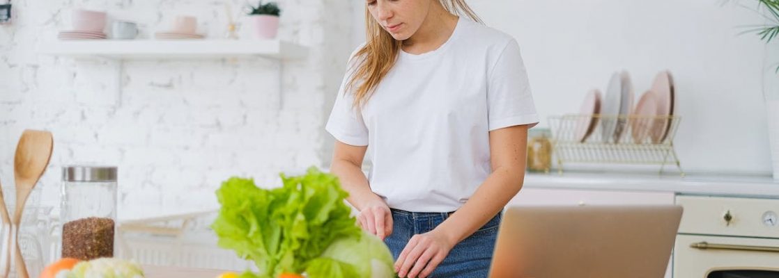 Woman in white t-shirt preparing fresh vegetables in a bright kitchen with a laptop.